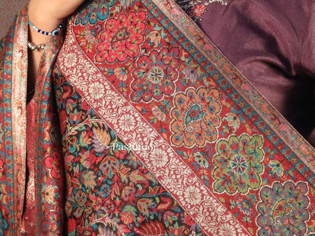 This image showcases an exquisite Pashnoor Shawl, featuring rich, intricate handwoven patterns inspired by traditional Kashmiri artistry. The shawl bursts with vibrant floral and paisley motifs in shades of red, teal, green, and gold, woven onto a deep, luxurious base. The detailed craftsmanship highlights the timeless beauty of pure pashmina, paired elegantly with a deep plum traditional outfit adorned with delicate embroidery. The overall look reflects heritage, luxury, and timeless grace — a true masterpiece from Pashnoor.
