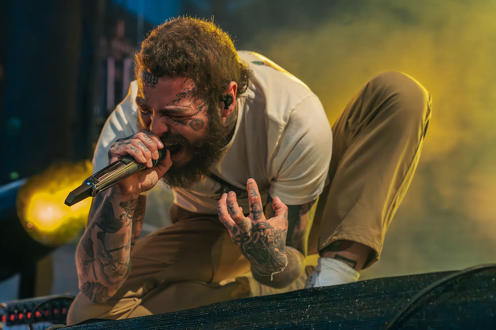 Post Malone leaning down clenching his fist upwards and holding a mic. Lights are flashing behind him