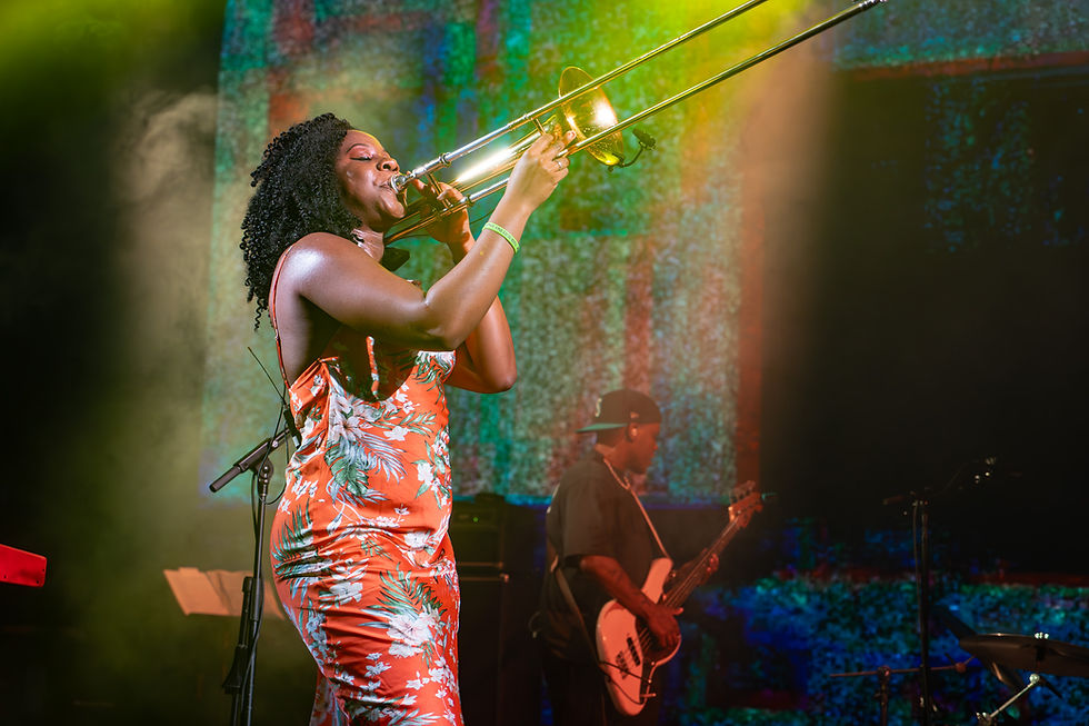 A Black female trombone player in a floral red dress performs on a lit stage