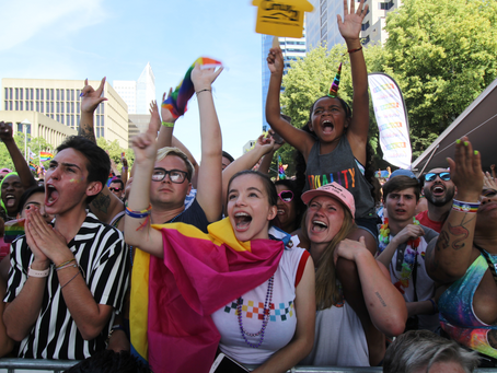 People at the Sacramento Pride Parade in 2023 gather around the main stage waiting for Lizzo, with kids on their parents shoulders screaming loudly, excited holding flags.