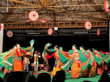 Women in colorful traditional attire dance energetically on stage with musicians in the background. Bright patterns and lively movements set a joyful mood.