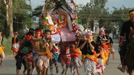 A group in colorful attire carries a decorated structure with a smiling figure during a vibrant street parade under a clear sky.