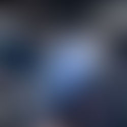 Two people sit by a small fire in a dim, rustic room with hanging meats and baskets. A cat stands near the fire. The mood is tranquil.