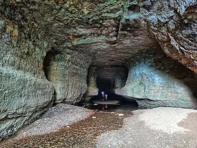 Two people with helmets explore a rocky cave with textured walls. The cave floor is wet, dimly lit, with earthy tones and shadows.