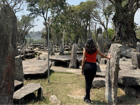 Woman in a red top walks through a forest of stone monoliths under a clear sky. Lush greenery surrounds the path, evoking serenity.