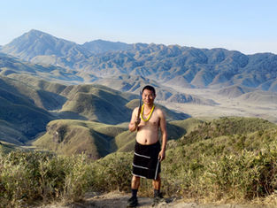 Man in traditional attire smiles, standing on a grassy hill with lush, rolling mountains in the background. Date on image: 2023.02.18.