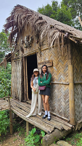 Two smiling women pose on a bamboo porch outside a thatched hut marked KHAWNBWL, surrounded by green trees.