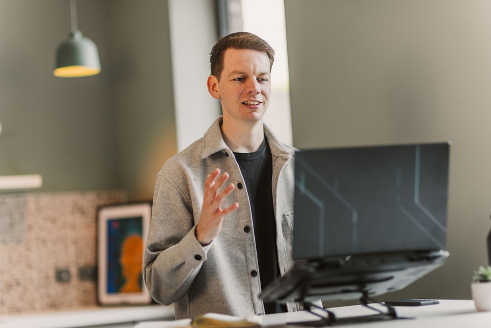 Person gesturing while speaking on a video call. Grey jacket, laptop on desk. Modern office with green walls and framed art.