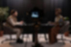 Two people sit across a table with microphones, engaged in conversation. Potted plants and a camera on a tripod are in the foreground.