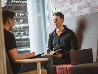 Two young men talk in a cozy office lounge, one taking notes with a notebook and laptop nearby, reversed window text in background.