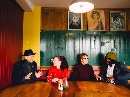 Four people sit around a wooden table, chatting in a warmly lit room with colourful walls and portraits. The mood is relaxed and sociable.