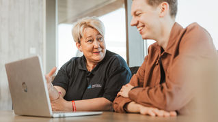 Two people sit at a table, engaged in a friendly discussion with a laptop open. The setting is bright and modern.