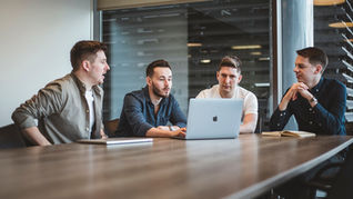 Four men sit around a table focused on a laptop. They're in a modern office with large windows, showing a collaborative discussion.