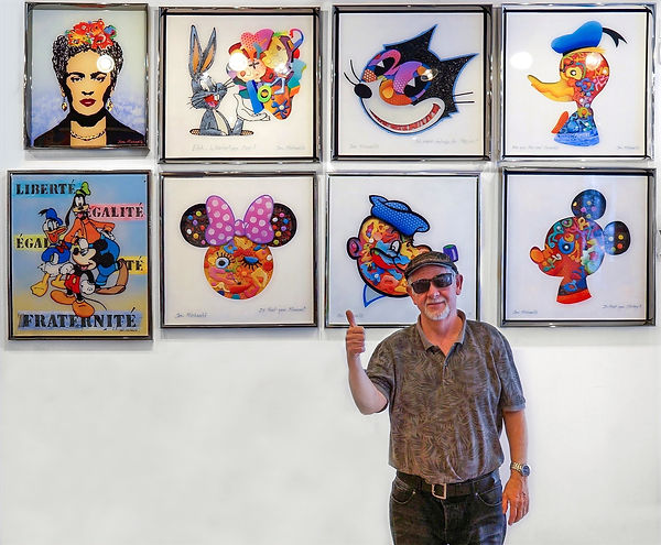 Jon smiling in a gallery in front of his paintings and gesturing a thumbs-up.