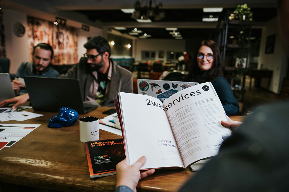 Business meeting scene with a woman in glasses smiling across from a man reading a service magazine, while two men beside her work on laptops and discuss ideas