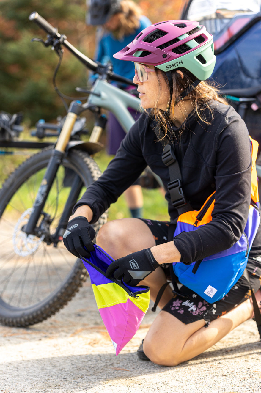 Cyclist kneels beside a bike, wearing pink helmet and colorful gear. Holds a purple, yellow, and pink pouch. Sunny outdoor setting.
