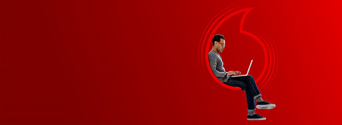 man sitting in a vodafone logo wearing a grey jumper and glasses whilst on his laptop
