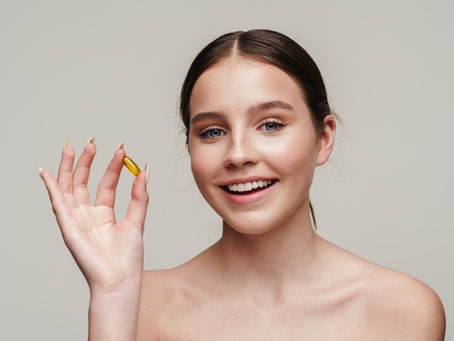 Mulher jovem com aparência saudável segurando cápsula de ômega 3 entre os dedos, sorrindo, em fundo neutro, representando suplementação e bem-estar.