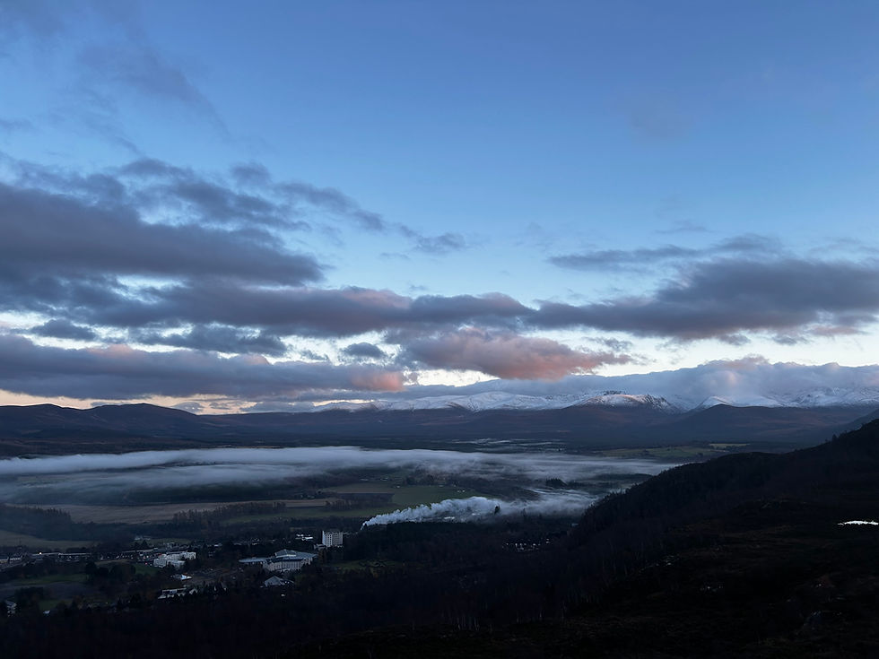Cairngorm Mountain landscape at dusk with snow-capped peaks, clouds, and light sky. Mist covers the valley. Smoke rises from a small settlement below.