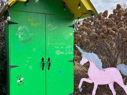 Bright green cabinet with positive messages, yellow roof, hanging bulbs, next to a pink unicorn cutout in a garden setting.