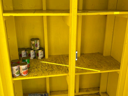 ellow wooden shelf with a few canned goods and boxes. Shelves mostly empty, casting shadows on a sunny day. Mood is sparse.