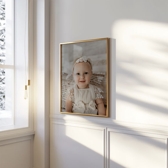 Framed wall portrait of a smiling baby girl wearing a lace romper and floral headband, beautifully displayed in a bright, sunlit room with modern white and gold décor. Luxury baby portrait by Elsiebel Photography in Burnsville, Minnesota — specializing in fine-art, light and airy newborn and milestone photography