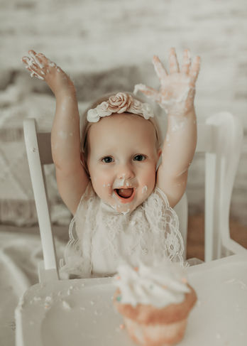 Happy baby girl raising her hands in excitement with cupcake in front of her. Light and airy cake smash portrait by Elsiebel Photography, Minneapolis metro.