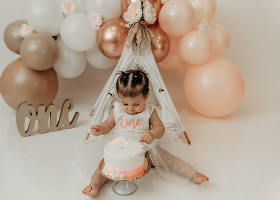 Baby girl sitting with her first birthday cake and balloon backdrop during a cake smash photography session.