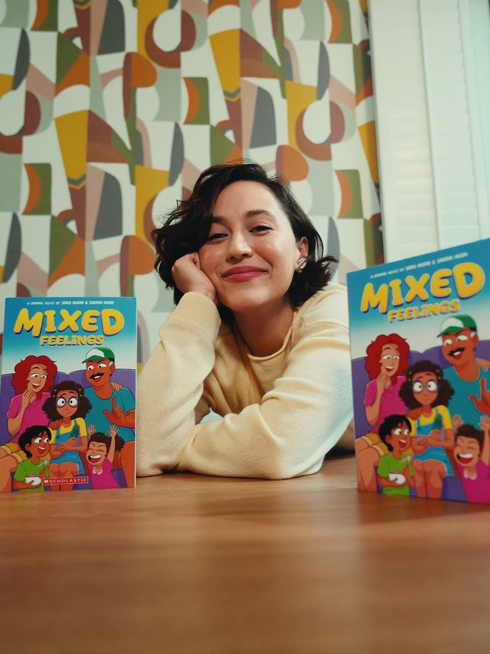 Sara Amini lounging on wooden floor with two  copies of her graphic novel"Mixed Feelings" 