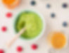 Green puree in a bowl with a spoon, surrounded by blueberries and raspberries on a white surface. Nearby, orange and yellow jars.