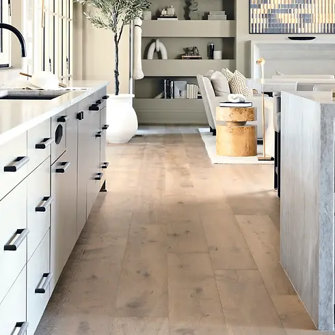 Modern kitchen with wide plank wood flooring, white cabinetry, and open living space in background.