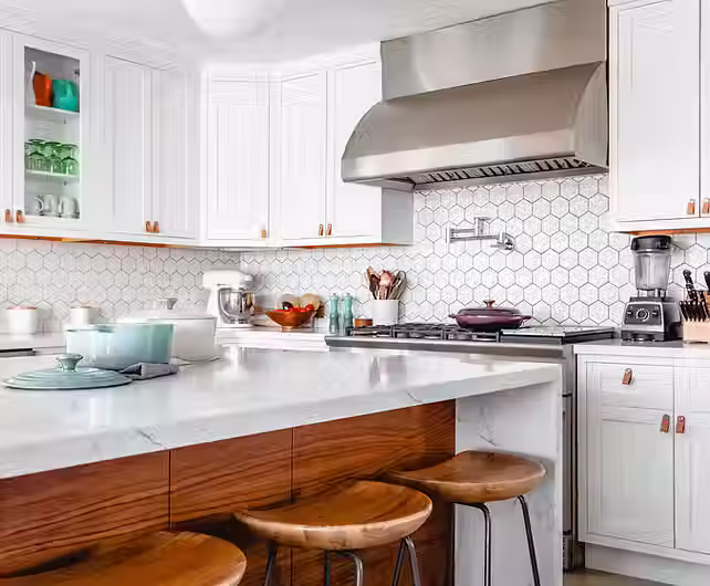 Modern kitchen features white cabinetry with leather pulls, a white marble waterfall island with wood-topped stools, and a distinctive hexagonal tile backsplash accented by a stainless steel range hood.