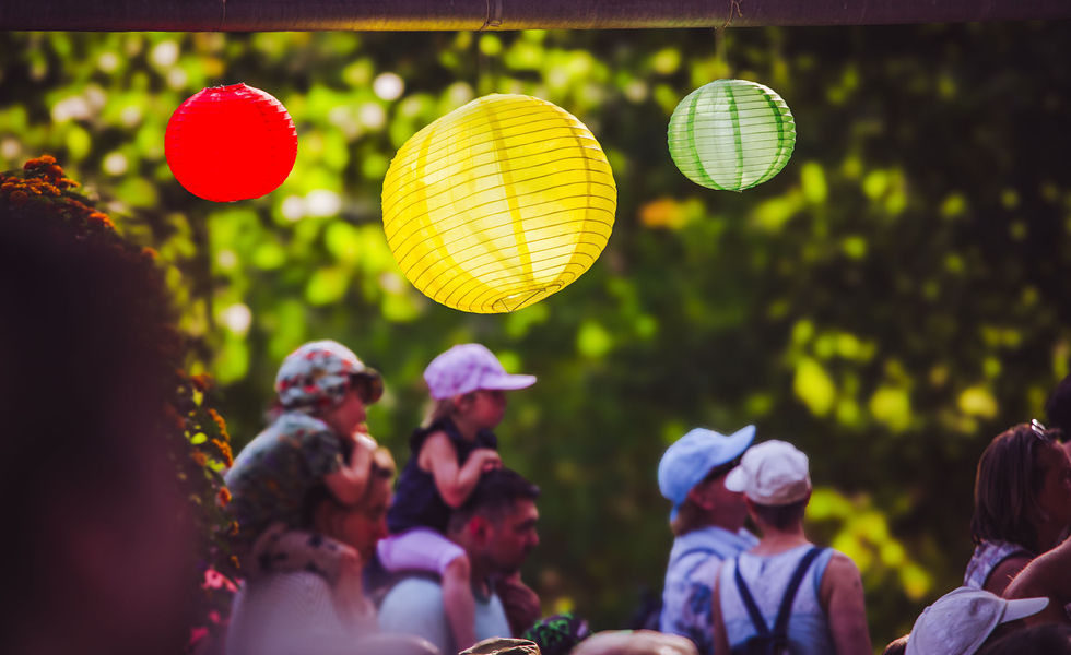 Im Vordergrund drei bunte Lampions an einer Pagoda. Im Hintergrund leicht unscharf ein paar Kleinkinder die auf den Schultern von Erwachsenen sitzen und Richtung einer Bühne schauen.