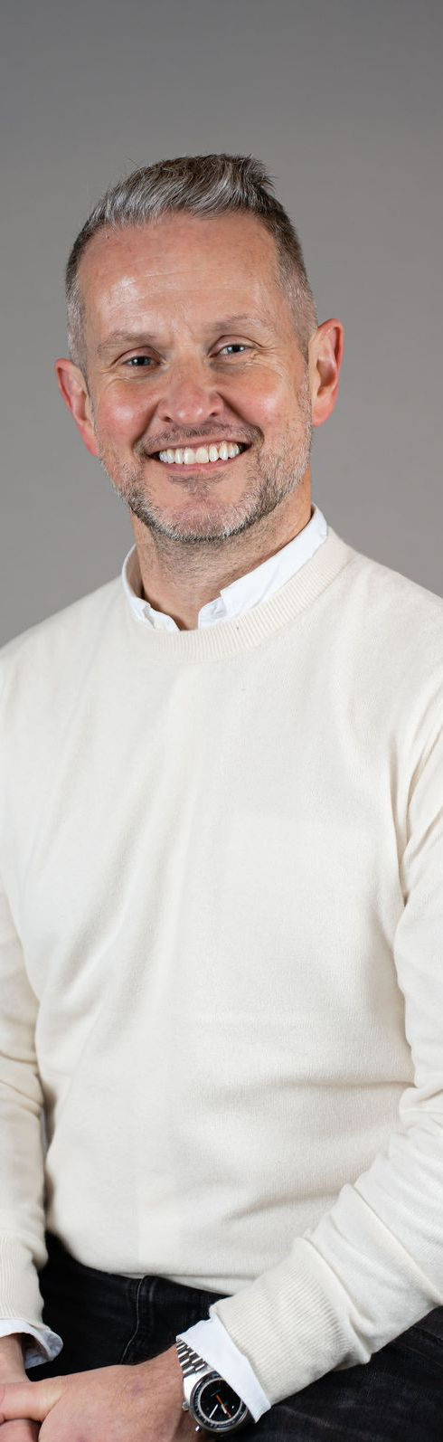 Photo of Relationship First Law's founder Andrew Magowan, sitting on a stool, smilig while wearing a white shirt under a cream jumper