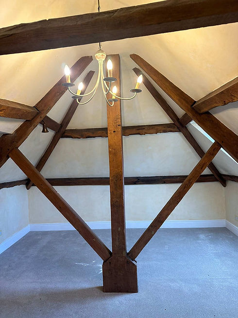 Vaulted attic room with rustic exposed wooden roof beams, carpeted floor, and a central candle-style chandelier.