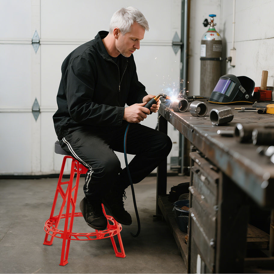 Man welding metal at a workbench, seated on a stool.