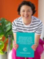 Sara holds a teal sign with text about growing ideas. Striped shirt, pink pants, orange wall, and green plant in the background.