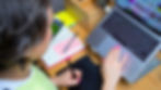 Sara uses her laptop and a tablet on a wooden desk. Open notebook with notes, pens, and colourful sticky notes nearby.