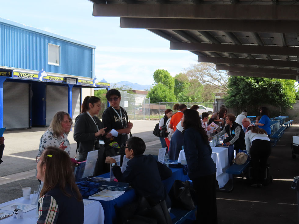 People gather at tables under a shelter, discussing materials. "HUSKY" signs on a blue building in the background. Sunny outdoor setting.