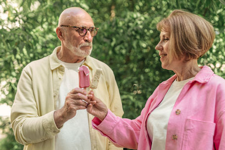 Older couple in pastel shirts share popsicles in a leafy park, smiling at each other in warm sunlight.