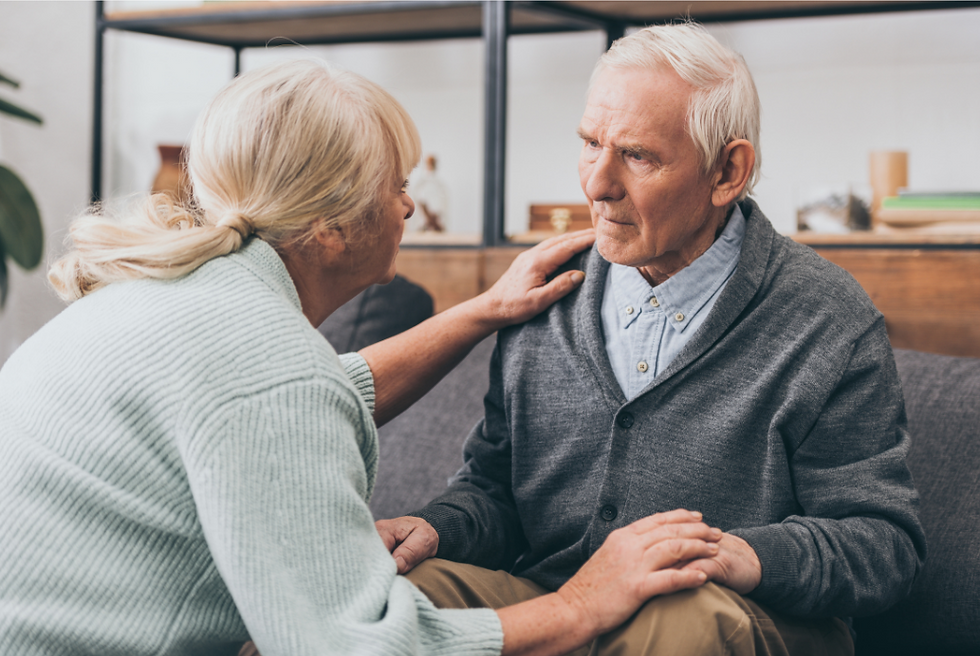 Elderly woman comforts an elderly man on a sofa. She wears a light sweater; he wears a gray cardigan. The setting is homey and calm.