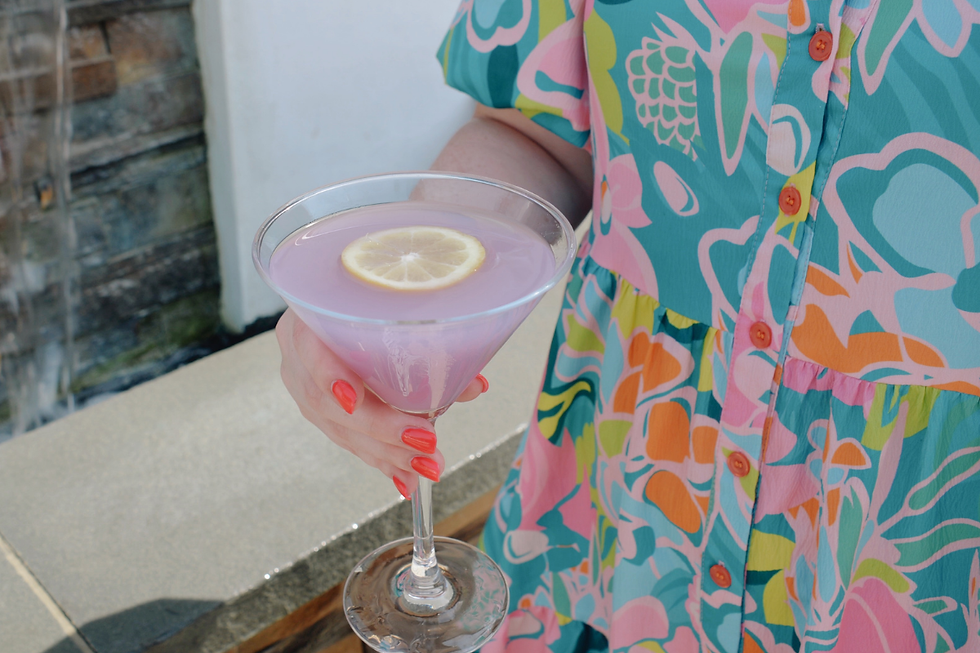 Person in a colorful floral dress holds a pink cocktail with a lemon slice beside a waterfall on the Farmer's Daughter Terrace.