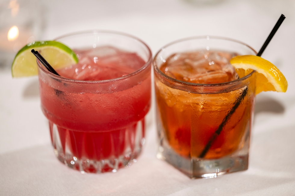 Two colorful cocktails with ice and straws, one red with lime and one amber with orange, on a white table in soft light at Normandy Farm.