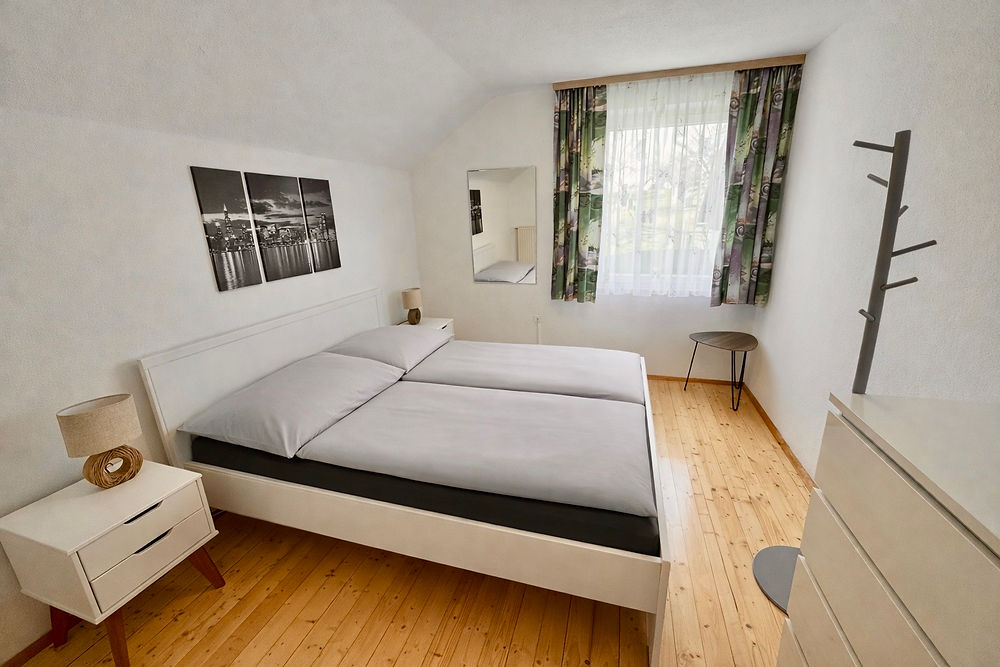 Schlafzimmer mit Doppelbett, grauer Bettwäsche, einem hellen Holzboden und weißen Wänden. 
