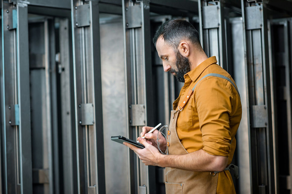 Man in an apron writes on a tablet with a stylus in an industrial setting, focused expression, metal beams in the background.