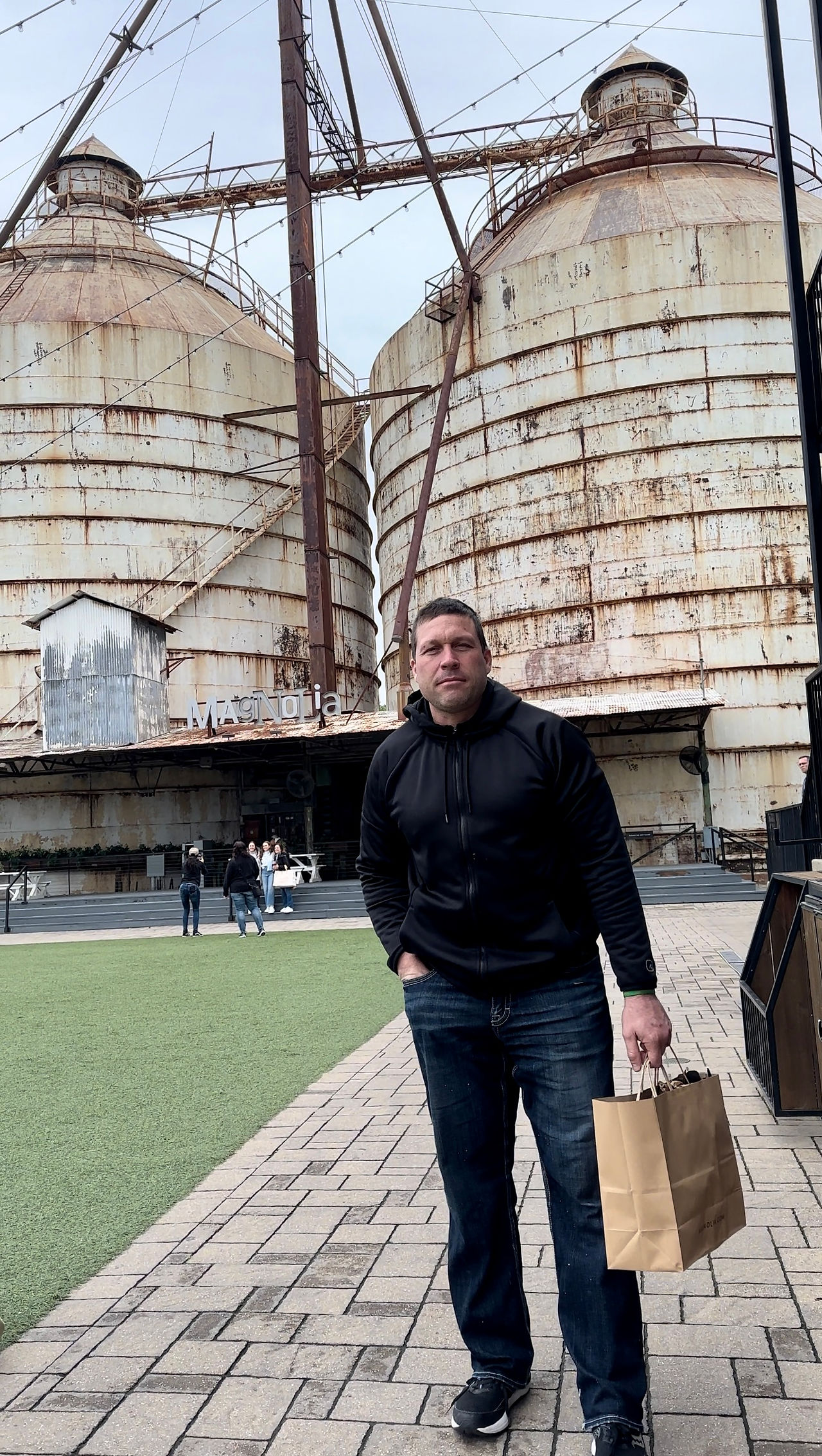 man in black sweatshirt holding.a shopping back in front of silos in magnolia waco Texas