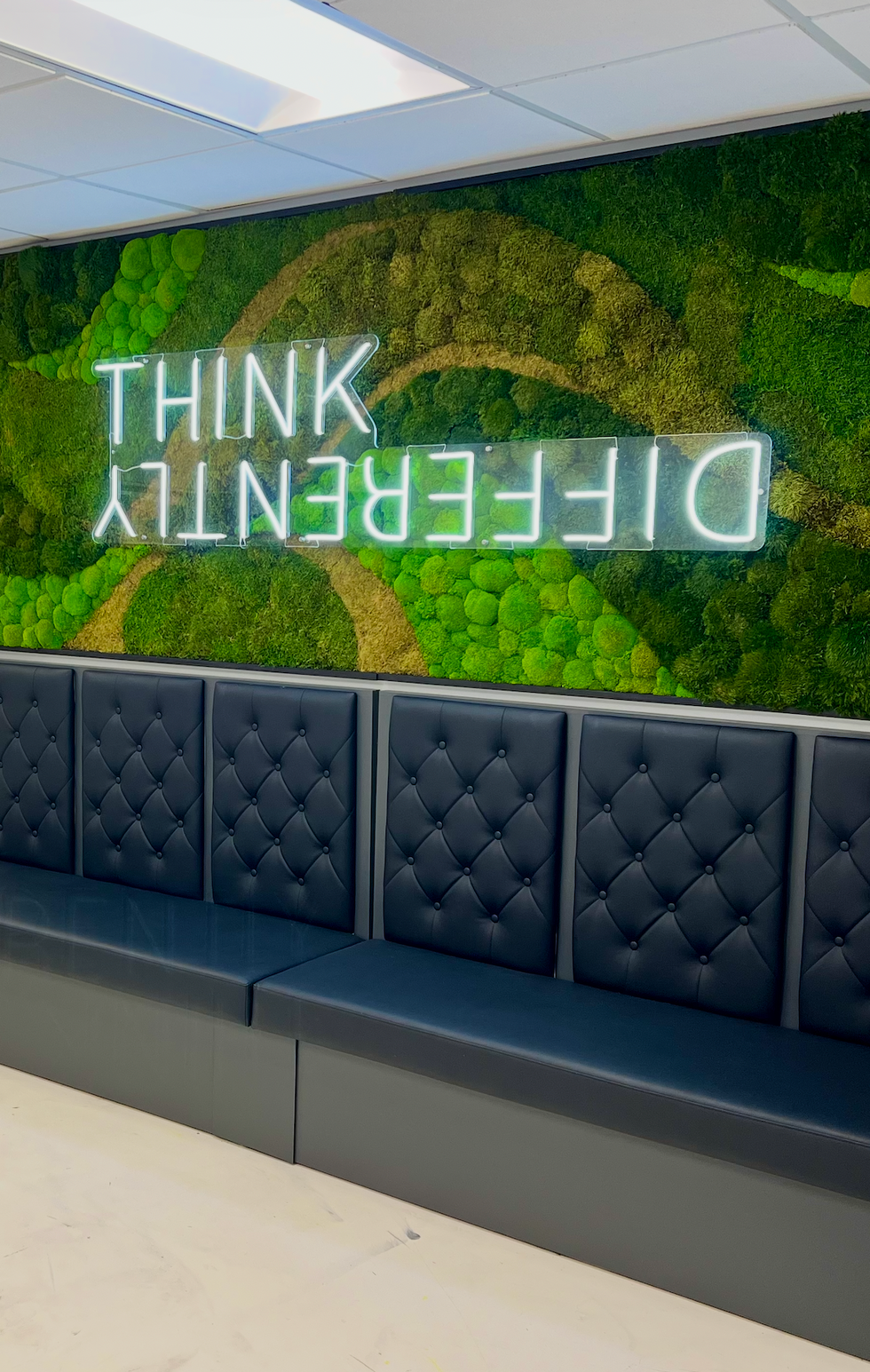Sleek breakroom corner in a modern office, showcasing a green moss wall with a bright LED motivational quote and dark quilted seating.