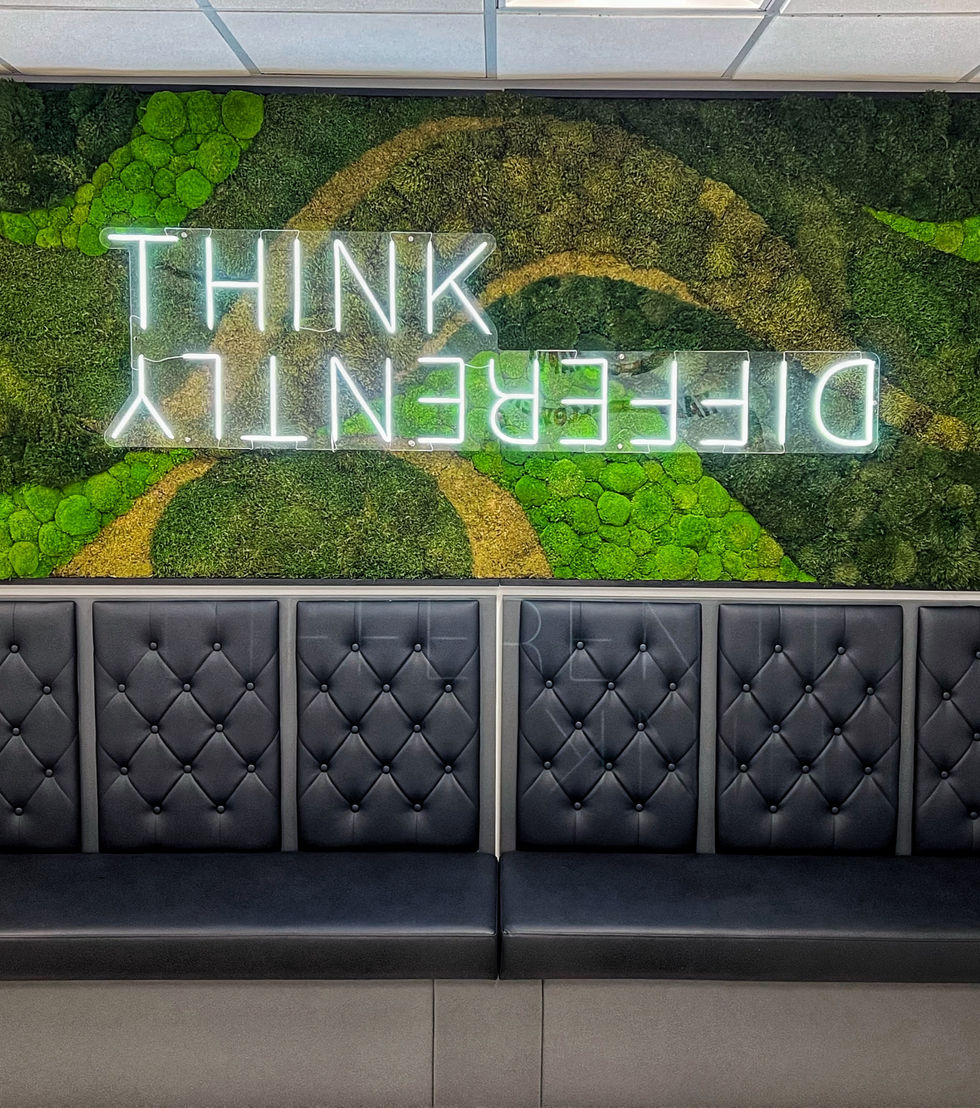 A modern office lounge with a green moss wall and illuminated “THINK DIFFERENTLY” LED sign above a row of dark tufted leather benches
