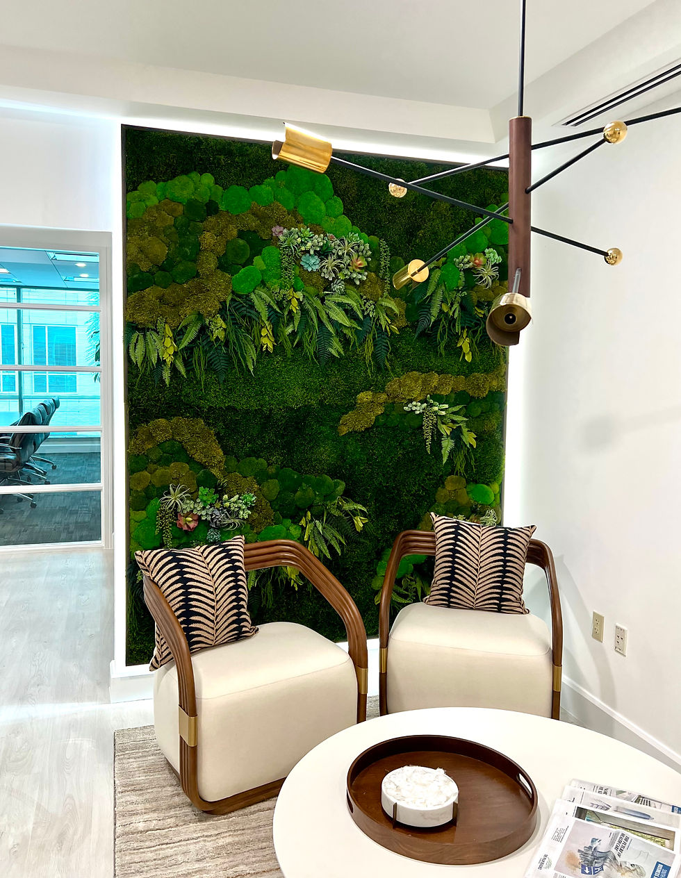 Modern room with a green plant wall, two beige chairs, patterned cushions, and a round table. A unique chandelier hangs above, creating a calm vibe.