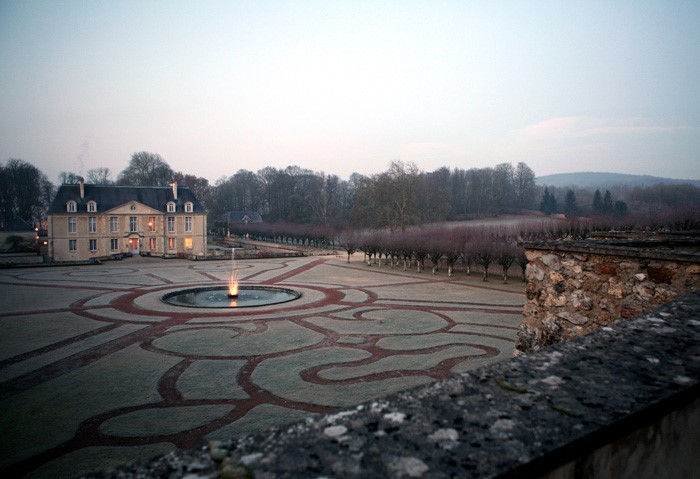 Symmetrical French formal garden with central fountain leading to Château de Louvois at dusk.